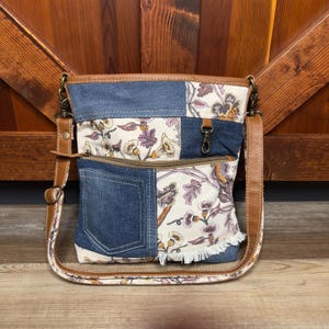 May include: A brown leather and denim patchwork crossbody bag with a floral print. The bag has a zippered pocket and a flap closure.
