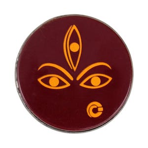 Op de afbeelding: Een ronde, kastanjebruine pin met een zilveren rand. De pin heeft een gestileerd oranje ontwerp van een gezicht met drie ogen en een gebogen symbool. Het ontwerp is gecentreerd op de pin.