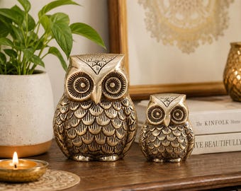 May include: Two metallic gold-toned owl figurines with detailed engraved feather patterns and circular eyes. The larger owl stands to the left of the smaller owl, both featuring pointed ear tufts and a smooth, polished finish on their beaks.