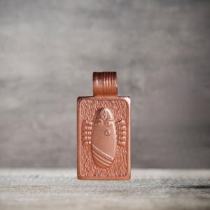 Op de afbeelding: Een koperen hanger met een rechthoekige vorm en een gedetailleerd ontwerp van een gestileerd gezicht met ogen, neus en mond. De hanger heeft een getextureerd oppervlak en een lus aan de bovenkant om een ​​ketting aan te bevestigen.