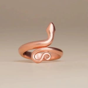 Op de afbeelding: Een ring van roségoudkleurig metaal in de vorm van een opgerolde slang. De band wikkelt zich in een cirkelvorm, waarbij de kop van de slang naar boven wijst en de staart aan de voorzijde naar binnen krult, wat zorgt voor een glad, gepolijst oppervlak.