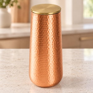 Puede incluir: Un recipiente cilíndrico de cobre con una superficie texturizada y martillada, junto con una tapa circular lisa de tono latón. El objeto presenta una forma cónica que se estrecha ligeramente hacia la parte superior, con un acabado metálico en tonos naranjas.