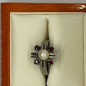 Puede incluir: Un anillo de plata con una perla central y cuatro piedras rojas. El anillo tiene un dise&ntilde;o en forma de cruz. El anillo se muestra en un joyero crema con marco de madera.