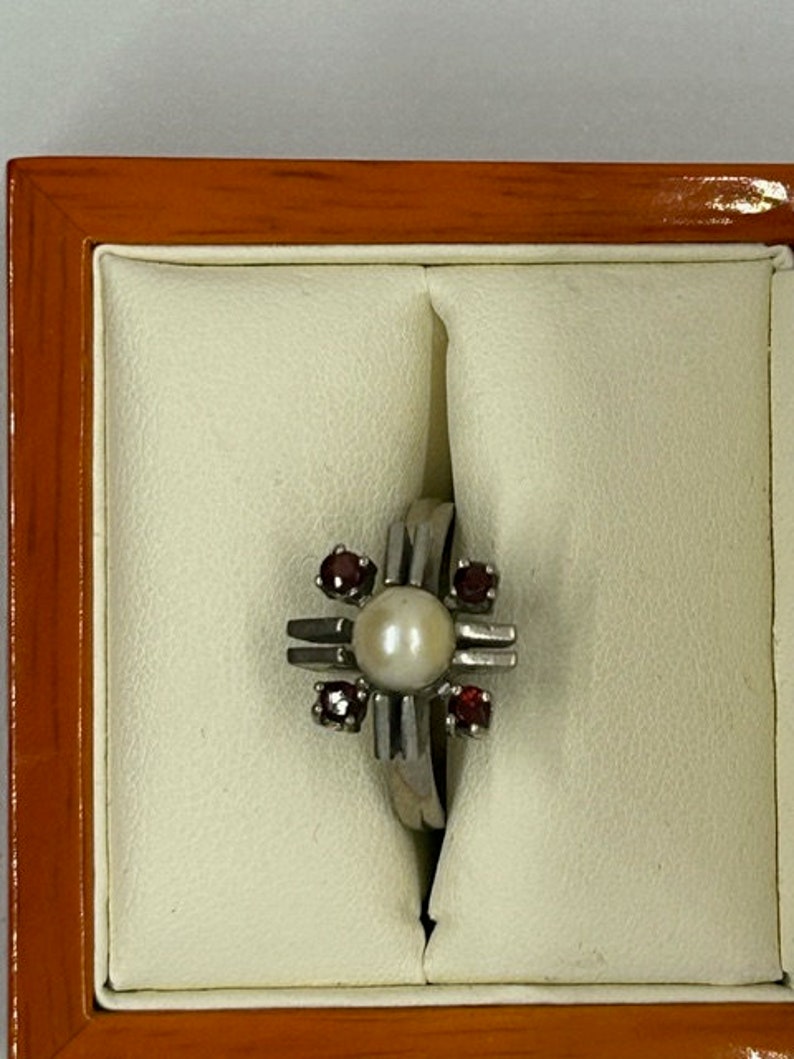 Puede incluir: Un anillo de plata con una perla central y cuatro piedras rojas. El anillo tiene un dise&ntilde;o en forma de cruz. El anillo se muestra en un joyero crema con marco de madera.