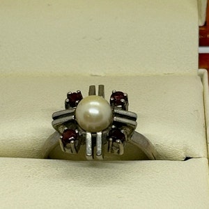 Puede incluir: Un anillo de plata con una perla central rodeada de cuatro piedras rojas m&aacute;s peque&ntilde;as. El anillo tiene un dise&ntilde;o geom&eacute;trico &uacute;nico con barras verticales y horizontales. El anillo se presenta en una caja de color claro.
