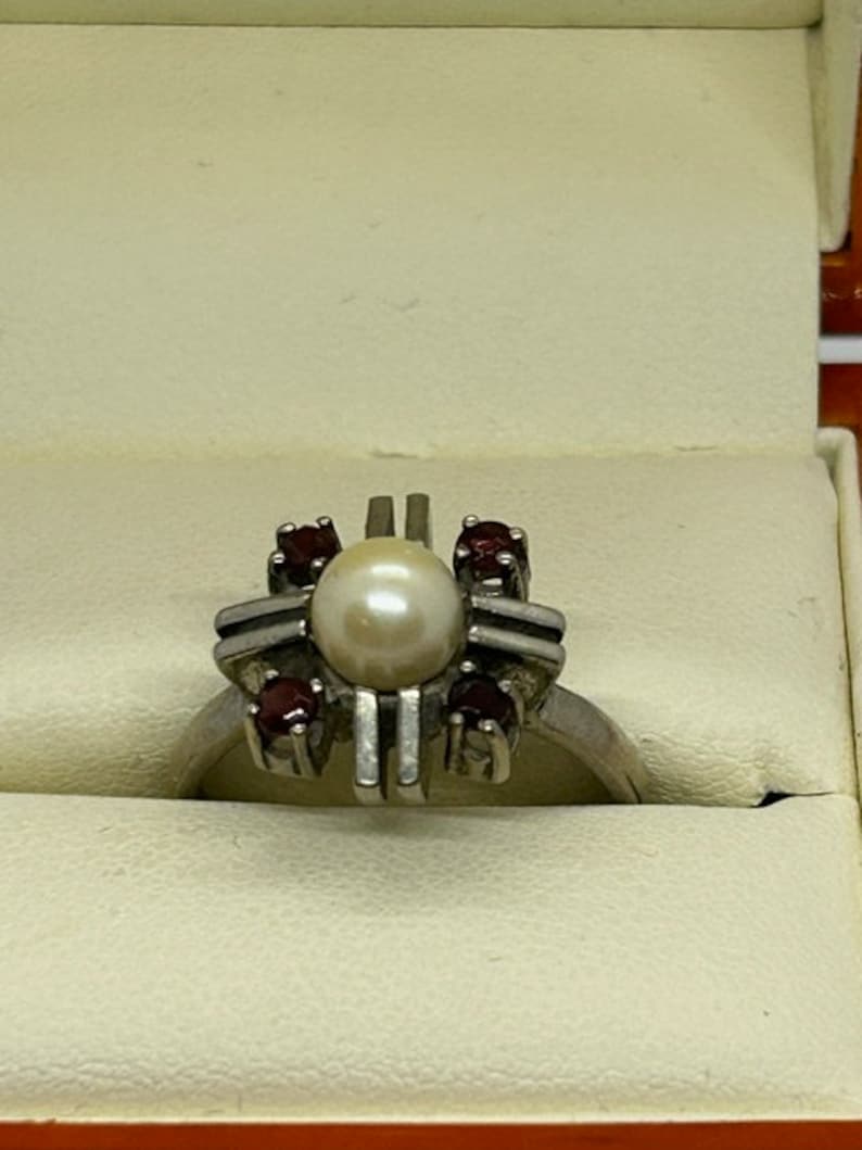 Puede incluir: Un anillo de plata con una perla central rodeada de cuatro piedras rojas m&aacute;s peque&ntilde;as. El anillo tiene un dise&ntilde;o geom&eacute;trico &uacute;nico con barras verticales y horizontales. El anillo se presenta en una caja de color claro.