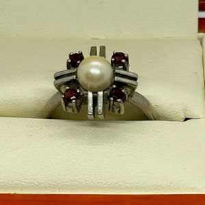 Puede incluir: Anillo de plata con una perla central y cuatro gemas rojas. El anillo tiene un dise&ntilde;o geom&eacute;trico &uacute;nico con barras verticales, sobre un fondo neutro.