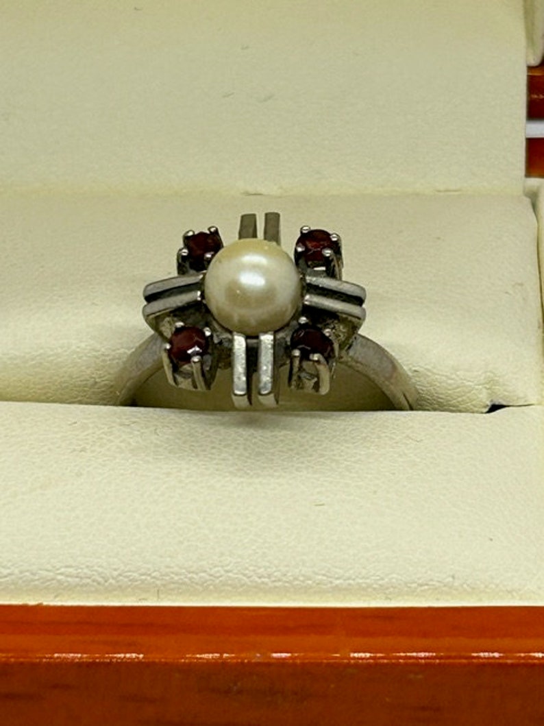 Puede incluir: Anillo de plata con una perla central y cuatro gemas rojas. El anillo tiene un dise&ntilde;o geom&eacute;trico &uacute;nico con barras verticales, sobre un fondo neutro.