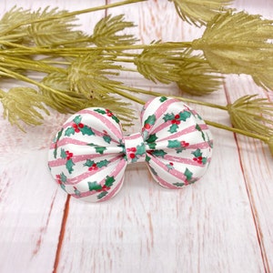 May include: A festive hair bow featuring a white background with pink stripes and polka dots. The bow is decorated with green holly leaves and red berries, perfect for the holiday season. The bow is a cute accessory.