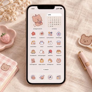 Op de afbeelding: Een smartphone met een aangepaste startscherminterface op een lichtbeige achtergrond, voorzien van een beer-illustratiewidget, een juni-kalender en diverse app-pictogrammen zoals foto's, camera, rekenmachine, instellingen en sociale media.