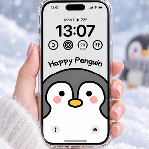 Pode incluir: Um smartphone com uma capa protetora transparente exibindo um papel de parede digital de um pinguim de desenho animado com bochechas rosas e bico amarelo. A tela mostra o texto Happy Penguin, a hora 13:07, data, clima e ícones de aplicativos.