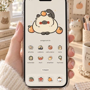 Op de afbeelding: Een smartphone met een doorzichtig hoesje waarop een aangepaste digitale interface wordt weergegeven. Het scherm bevat een centrale illustratie van een ronde kat met sinaasappels, de tekst widgetsmith en diverse app-pictogrammen met de labels photos, camera, calculator, note, calendar, mail, weather en factime.