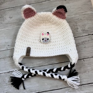 May include: White crocheted hat with cat ears and a cat face applique. The hat has black and white braided ties.
