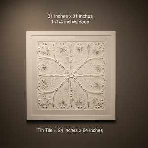 May include: A white framed artwork featuring a white embossed tin tile with a floral design. The artwork measures 31 inches by 31 inches and is 1 1/4 inches deep. The tin tile itself measures 24 inches by 24 inches.
