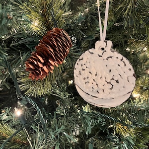 May include: A white ceramic Christmas bauble with a textured surface hangs on a green Christmas tree with brown pine cones. The bauble is decorated with a white and black speckled pattern.