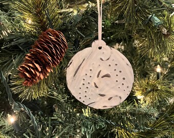 May include: White painted ornament with a black and white design hanging on a green Christmas tree with brown pine cones.