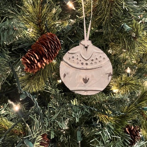 May include: A white ceramic Christmas bauble with a textured surface and a snowflake design. The bauble is hanging on a green Christmas tree.