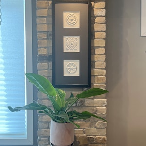 May include: A framed artwork with three white, textured relief tiles, each with a different geometric design. The artwork is set against a dark grey background and framed in black. A potted plant with large green leaves is positioned below.