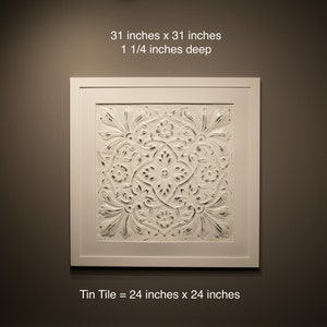 May include: A white framed wall art piece with a raised embossed design in the centre. The art piece measures 31 inches by 31 inches and is 1 1/4 inches deep. The text "Tin Tile = 24 inches x 24 inches" is below the framed art piece.