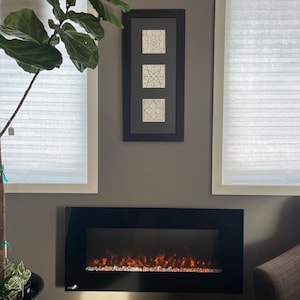 May include: A black electric fireplace with a realistic flame effect is mounted on a wall in a living room. The fireplace is surrounded by white trim and a grey wall. A large green plant is in the corner of the room.