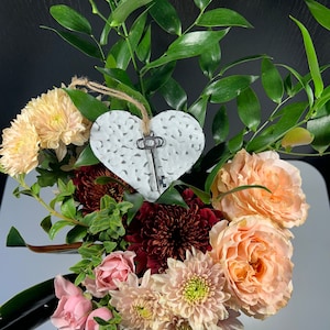 May include: A bouquet of pink, peach, and burgundy flowers with a white heart-shaped ornament featuring a key design. The flowers are arranged in a vase.
