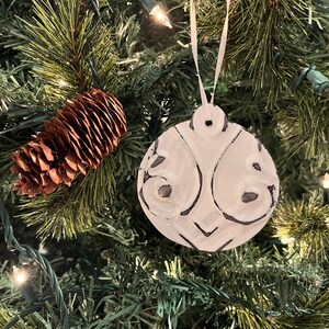 Könnte beinhalten: Weißer Keramik-Weihnachtsbaumschmuck mit einem dekorativen Design. Der Schmuck hängt an einem grünen Weihnachtsbaum mit braunen Tannenzapfen.
