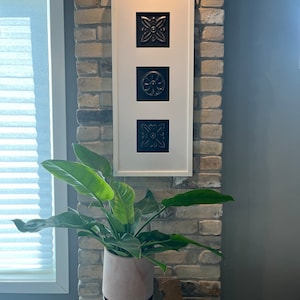 May include: A white framed wall art piece with three black and gold decorative tiles. The art piece is hanging on a brick wall next to a potted green plant.