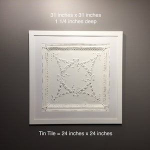 May include: A white framed artwork featuring a white tin tile with a decorative embossed design. The tile measures 24 inches by 24 inches and is framed to a size of 31 inches by 31 inches. The frame is 1 1/4 inches deep.