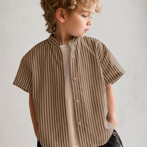 May include: A short-sleeved, button-down shirt with vertical brown and beige stripes. The shirt has a mandarin collar and is worn over a light-colored undershirt. The child is wearing black pants.