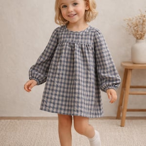 May include: A young child wearing a blue and beige checkered dress with long sleeves and a round neckline. The dress is knee-length. The child is also wearing white socks and a white hair bow.