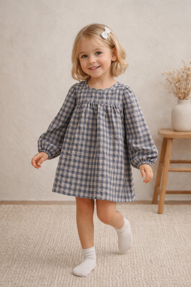 May include: A young child wearing a blue and beige checkered dress with long sleeves and a round neckline. The dress is knee-length. The child is also wearing white socks and a white hair bow.