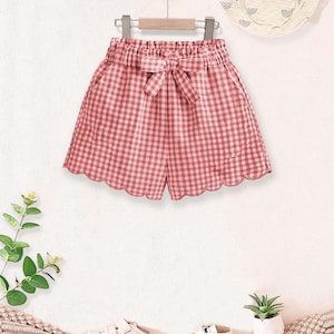 May include: Pink and white gingham shorts with a bow detail, scalloped hem, and elastic waistband. A matching sun hat with a rainbow design and white sneakers are also visible. The shorts are hanging on a wooden hanger.