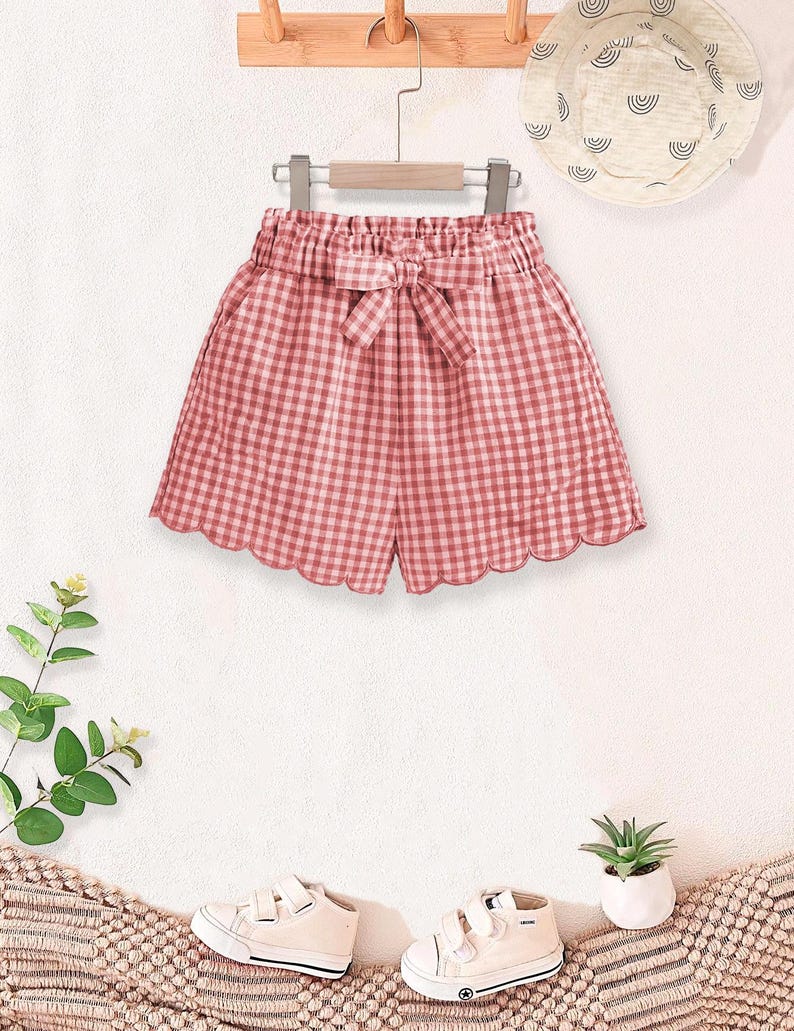 May include: Pink and white gingham shorts with a bow detail, scalloped hem, and elastic waistband. A matching sun hat with a rainbow design and white sneakers are also visible. The shorts are hanging on a wooden hanger.