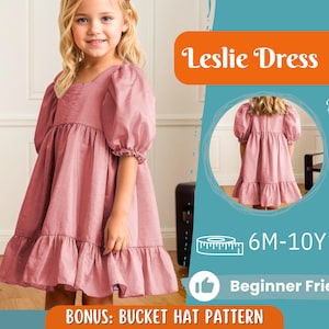 May include: A young girl wearing a dusty rose-colored dress with a square neckline and ruffled hem. The dress has short, puffy sleeves. The image includes the text "Leslie Dress", "6M-10Y", "Beginner Friendly", and "BONUS: BUCKET HAT PATTERN".