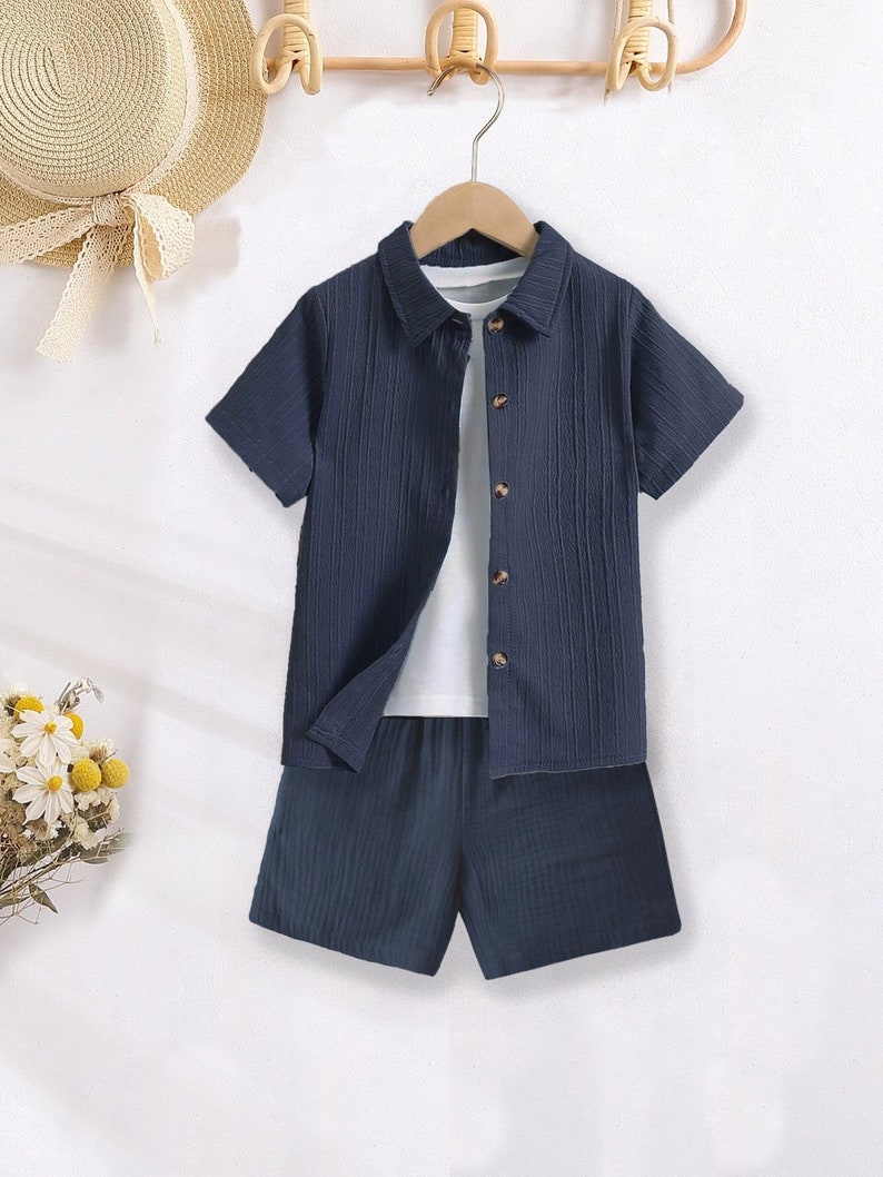 K&ouml;nnte beinhalten: Ein marineblaues kurz&auml;rmeliges Hemd und Shorts-Set mit wei&szlig;em Unterhemd. Das Hemd hat eine Knopfleiste und ein strukturiertes vertikales Streifenmuster. Die Shorts sind in passendem Marineblau gehalten.
