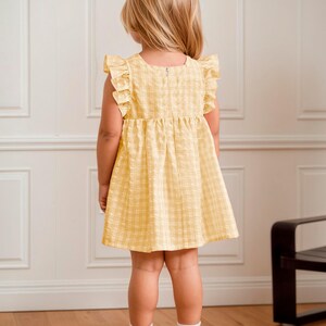 May include: A yellow plaid dress with ruffled sleeves. The dress is worn by a young child, who is facing away from the camera. The dress has a simple, classic design, perfect for a summer day. The child is wearing white socks and shoes.
