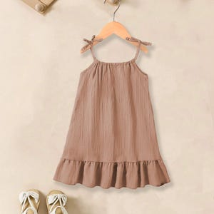 May include: A light brown, sleeveless dress with tie straps and a ruffled hem, hanging on a wooden hanger. Two pairs of white sandals with bow accents are at the bottom of the image.