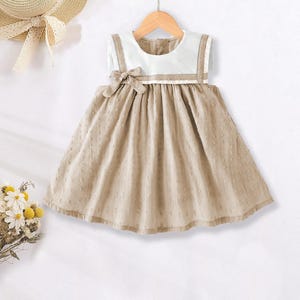 May include: A beige baby dress with a white collar and straps, hanging on a wooden hanger. The dress features a bow at the waist and a dotted pattern on the skirt. A straw hat with a ribbon is visible in the background.