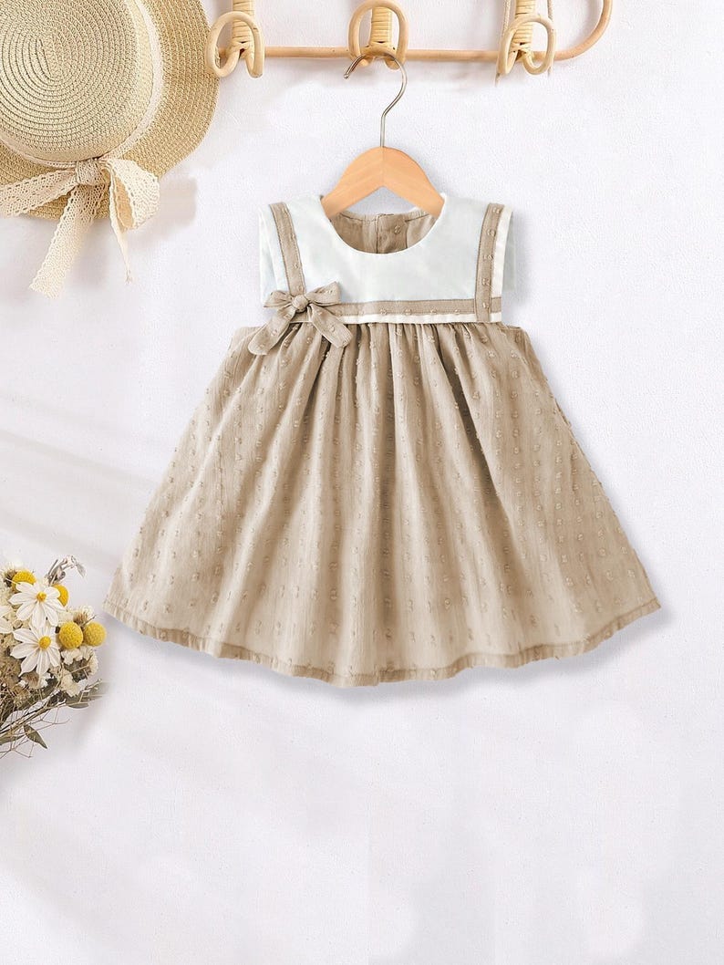 May include: A beige baby dress with a white collar and straps, hanging on a wooden hanger. The dress features a bow at the waist and a dotted pattern on the skirt. A straw hat with a ribbon is visible in the background.