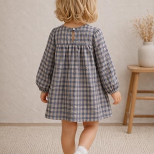 May include: A little girl wearing a blue and tan plaid dress with long sleeves and a keyhole back. The dress is knee-length. She is wearing white socks and a white bow in her hair. The background includes a wooden stool and a vase with dried flowers.