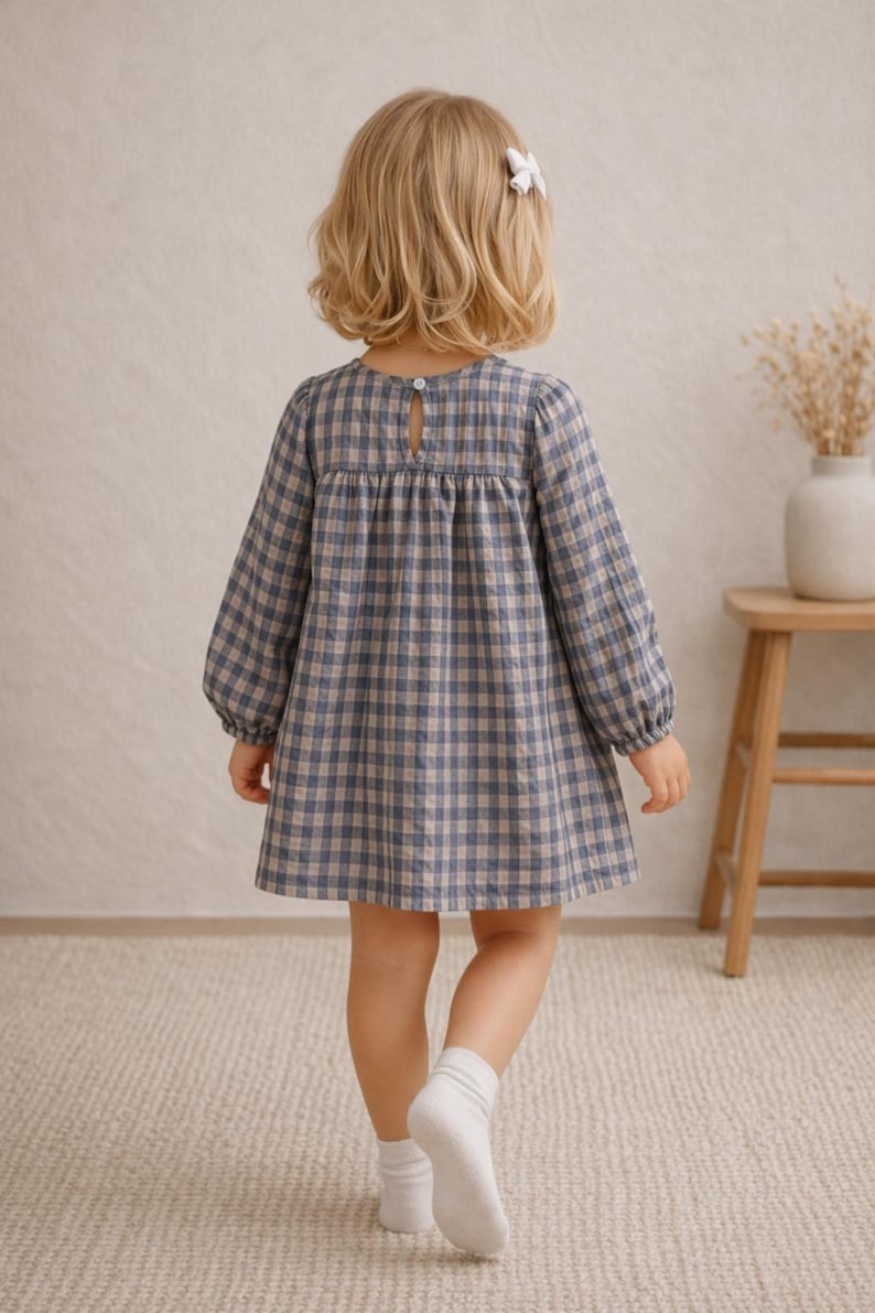 May include: A little girl wearing a blue and tan plaid dress with long sleeves and a keyhole back. The dress is knee-length. She is wearing white socks and a white bow in her hair. The background includes a wooden stool and a vase with dried flowers.