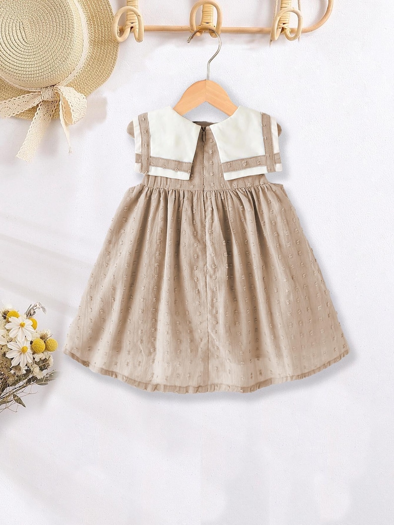 May include: A tan dress with a white collar and a small dot pattern. The dress is hanging on a wooden hanger. A straw hat with a ribbon is visible in the background.