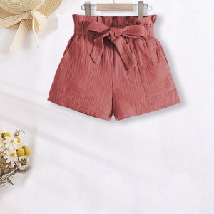 Puede incluir: Pantalones cortos de color rosa polvoriento con cintura alta fruncida y lazo de tela a juego. Los pantalones cortos tienen dos bolsillos delanteros y cuelgan de una percha de madera. Un sombrero de paja con una cinta es visible en el fondo.