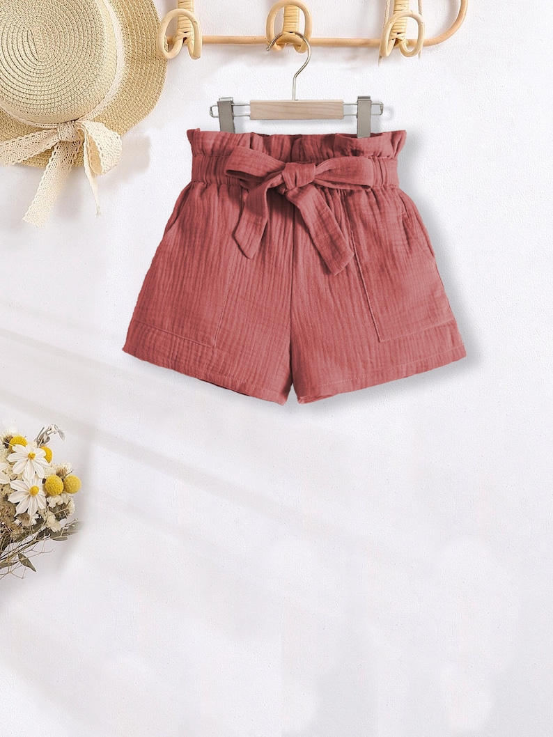 Puede incluir: Pantalones cortos de color rosa polvoriento con cintura alta fruncida y lazo de tela a juego. Los pantalones cortos tienen dos bolsillos delanteros y cuelgan de una percha de madera. Un sombrero de paja con una cinta es visible en el fondo.