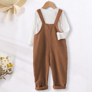 May include: A brown corduroy overall with a white long-sleeved shirt underneath. The overall has adjustable straps and a button closure.