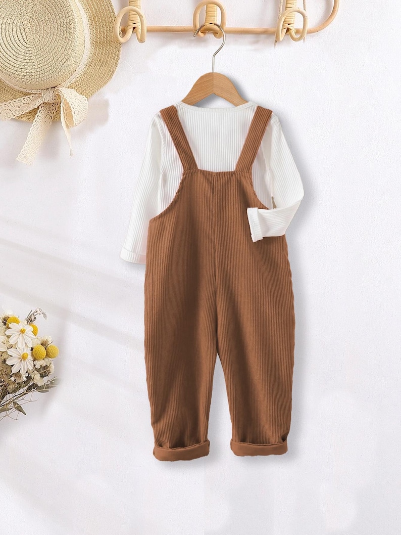 May include: A brown corduroy overall with a white long-sleeved shirt underneath. The overall has adjustable straps and a button closure.
