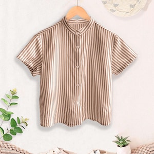 May include: A short-sleeved, button-down shirt with vertical brown and white stripes hangs on a wooden hanger. The shirt has a mandarin collar. A white sun hat with a black pattern hangs above the shirt. White sneakers and a small plant are below.