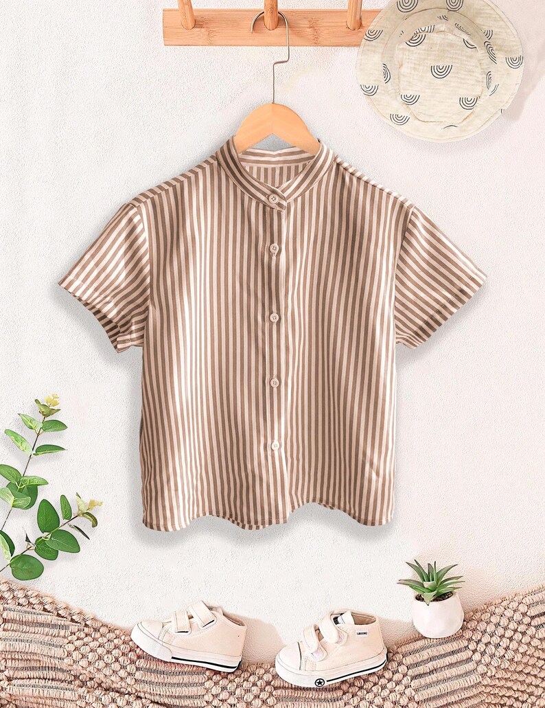 May include: A short-sleeved, button-down shirt with vertical brown and white stripes hangs on a wooden hanger. The shirt has a mandarin collar. A white sun hat with a black pattern hangs above the shirt. White sneakers and a small plant are below.