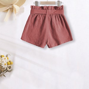 Puede incluir: Pantalones cortos de color rosa empolvado con cintura alta y cintur&oacute;n a juego, colgados de una percha de madera. Tambi&eacute;n se ve un sombrero de paja con una cinta blanca y un ramo de flores secas. La tela es texturizada.