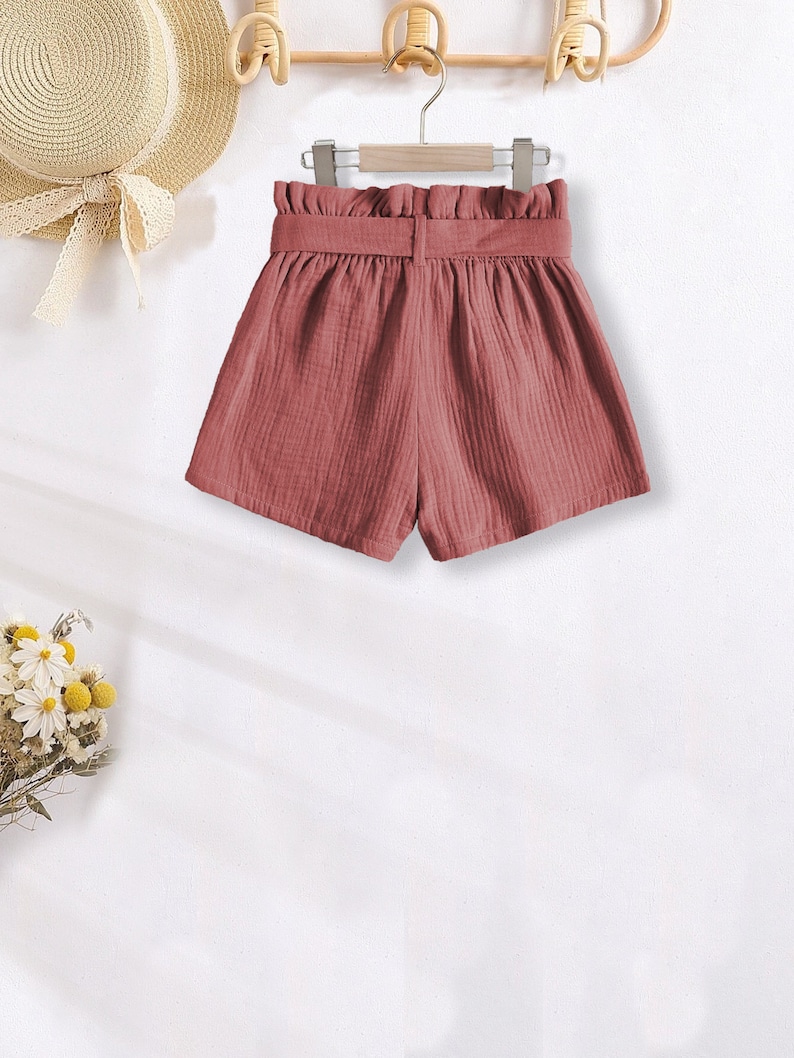 Puede incluir: Pantalones cortos de color rosa empolvado con cintura alta y cintur&oacute;n a juego, colgados de una percha de madera. Tambi&eacute;n se ve un sombrero de paja con una cinta blanca y un ramo de flores secas. La tela es texturizada.